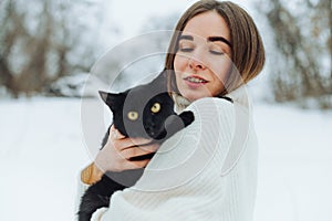 Positive woman in a sweater on a winter walk with a black cat in his hands standing on a snowy street