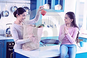 Positive woman taking red paper from paper bag and showing to daughter