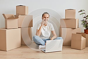 Positive woman wearing white T-shirt sitting on the floor near cardboard boxes with personal stuff, talking phone, looking at