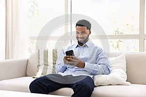 Positive young African man typing on cellphone, relaxing on couch