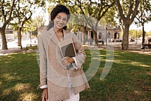 Positive young african woman looking into camera stands in park with folders and tablet.