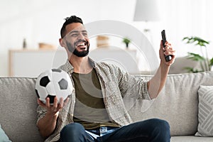 Positive young Arab guy watching soccer game on TV, holding remote control and ball at home