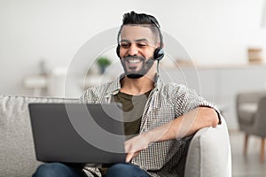Positive young Arab man wearing headphones, having online meeting on laptop at home