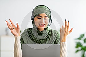Positive young Arab woman in hijab wearing headphones, having online meeting on laptop at home