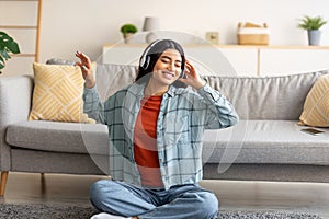 Positive young Arab woman sitting on floor with headphones, listening to music or audio book at home