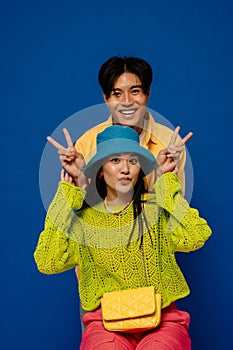 Positive asian couple showing peace sign while posing isolated over blue wall