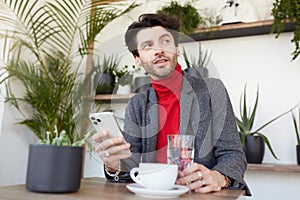 Positive young attractive brown haired unshaved male looking pensively aside while sitting over cafe interior in trendy formal