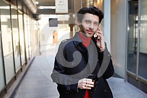 Positive young attractive brown haired unshaved male in trendy clothes holding takeaway coffee while making call with his