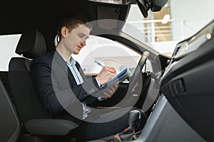 Positive young auto salesman sitting inside new car, making checkup, taking notes during test drive at dealership