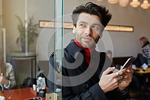 Positive young beautiful brown haired bearded guy keeping mobile phone in raised hands and looking pensively in window, posing