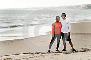 Positive young black couple in sportswear and wireless headphones hugs relax after running on sea beach