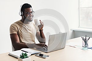 Positive young black guy in headphones drinking coffee, using laptop at desk in home office, copy space