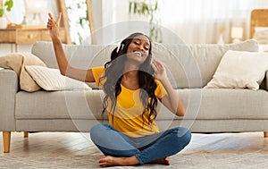 Positive young black woman in headphones listening to music on floor in living room