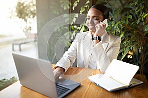 Positive young black woman in white suit at table typing on computer, calls by phone in cafe with plants interior