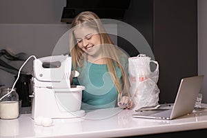 Positive young blonde woman standing at kitchen table, cooking dinner, watching culinary TV show online or cooking tutorial on