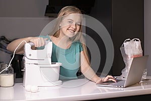 Positive young blonde woman standing at kitchen table, cooking dinner, watching culinary TV show online or cooking tutorial on