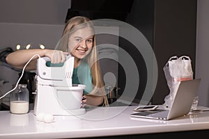 Positive young blonde woman standing at kitchen table, cooking dinner, watching culinary TV show online or cooking tutorial on