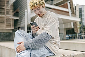 Positive young caucasian blond man using smartphone sits near library.