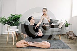 Positive young Caucasian couple having online yoga class at home, using laptop, sitting in lotus pose. Pretty lady