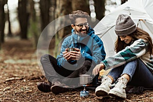 Positive young caucasian family in jackets sitting, relaxing near tent, prepare hot drink, enjoy adventure