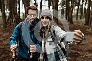 Positive young caucasian family take photo on phone, selfie, enjoy adventure, free time, walk in cold forest in autumn