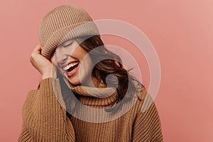 Positive young caucasian girl closes eyes and smiles teeth pulling hat over face on pink background.