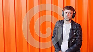 Positive young caucasian man is standing in the music headphones and looking at the camera on an orange background