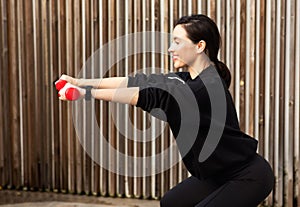 Positive young caucasian slim woman in sportswear doing exercise with dumbbells with hands and squat