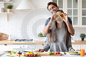 Positive young couple in love having fun while cooking in kitchen at home, making eyeglasses from bell pepper slices. Enjoying