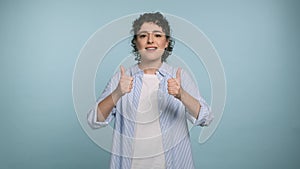 Positive young curly woman showing thumbs up, gazes happy at camera and enjoying posing on blue background. Positive