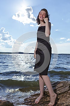 Positive Young Happy Smiling Caucasian Brunette Girl Posing in Black Dress On Stone At Sea During Sunny Day Outdoors