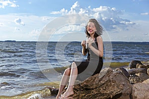 Positive Young Happy Smiling Caucasian Brunette Girl Posing in Black Dress On Stone At Sea During Sunny Day Outdoors