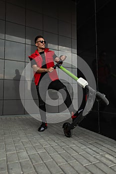 Positive young hipster man with hairstyle in stylish denim red-black clothes in sunglasses poses with a modern electric scooter