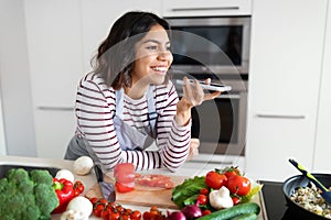 Positive young hispanic woman recording voice message while cooking