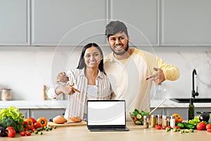 Positive young indian couple cooking at home, showing laptop