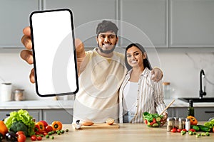 Positive young indian couple cooking at home, showing phone