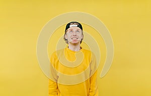 Positive young man in a cap and sweatshirt with a radiant face looking up on a yellow background