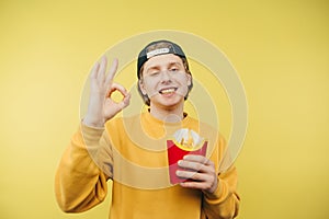 Positive young man in casual clothes holds french fries in his hands and shows OK gesture to the camera with a smile on his face