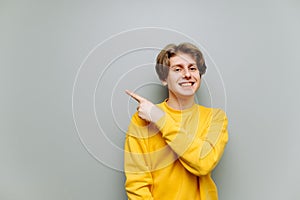 Positive young man looking at camera and pointing finger to the side at copy space with smile on face, isolated on gray background