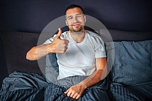 Positive young man sitting on bed and looks on camera. He smiles and shows big thumbs up. Guy is in bedroom.