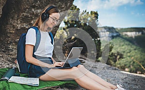 Positive young millennial girl with headphones sitting outdoors in mountains working from distance using laptop, beautiful tourist