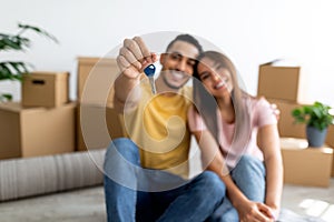 Positive young multiracial couple showing house key, sitting on floor of new home, selective focus
