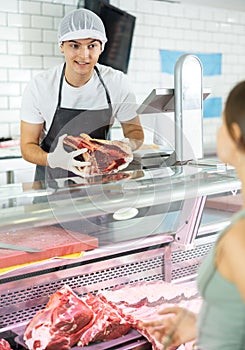 Positive young salesman demonstrating piece of meat to purchaser in butcher shop