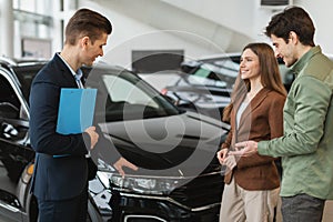 Positive young spouses speaking to salesman about purchasing new auto at dealership shop. Car purchase or rental