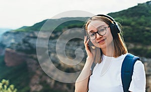 Positive young traveler with long blonde hair and hipster glasses listens to favorite music in wireless headphones smiles