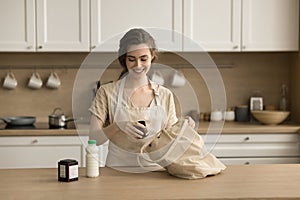 Positive young woman in apron unpacking shopping bag in kitchen