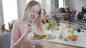 Positive young woman chewing tasty organic salad and smiling at camera. Portrait of joyful blond lady eating healthful