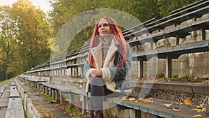 Positive young woman with dreadlocks looking away, smiling, sitting on old shabby bench of street bleacher in golden