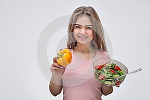 Positive young woman hold bowl with salad and yellow pepper in hands. She look on camer and smile. Isolated on grey