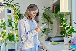Positive young woman holding smartphone in hands, looking at screen, in home
